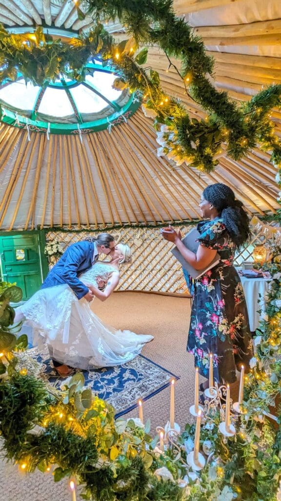 The celebrant applauds while the groom holds his bride in a cheeky embrace to enjoy their first kiss as husband and wife during their private elopement wedding ceremony in the yurt.