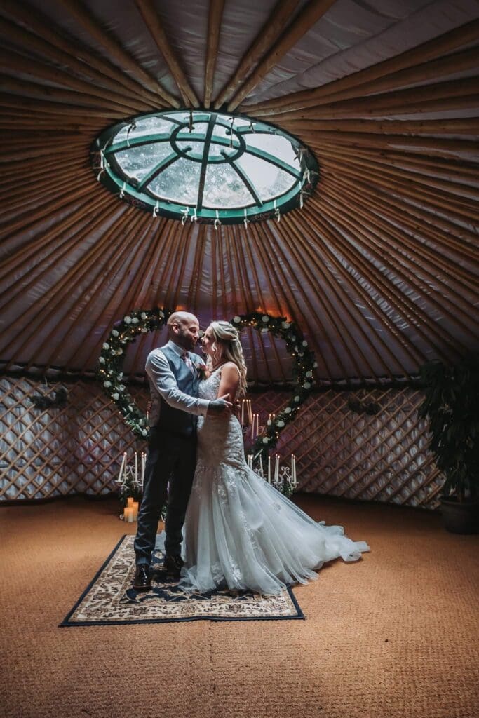 A bride and groom hold each other in an embrace in the woodland wedding yurt with the large heart shaped wedding arch backdrop for intimate elopement weddings in Yorkshire.