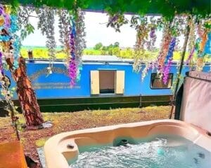 Luxury narrowboat moored beside a bubbling hot tub on a patio deck, surrounded by hanging flowers and greenery. A romantic waterside retreat in Yorkshire.