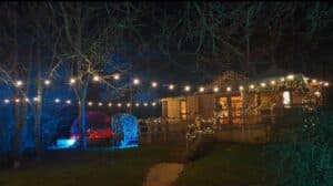Romantic woodland lodge at night with warm string lights, illuminated trees, and a glowing stargazing cinema dome, creating a cosy luxury escape at Hawthorn Hideaway. The perfect alternative to glamping with a hot tub.