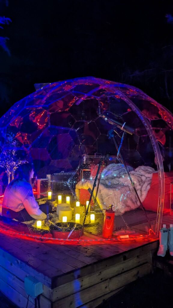 A cosy dome at night, lit by candlelight and blues and reds of soothing colour changing LED's. Inside is set for a cosy night time sound bath and the sofa and fluffy throws helps couples relax. A great way to end the day during a romantic getaway at Hawthorn Hideaway.